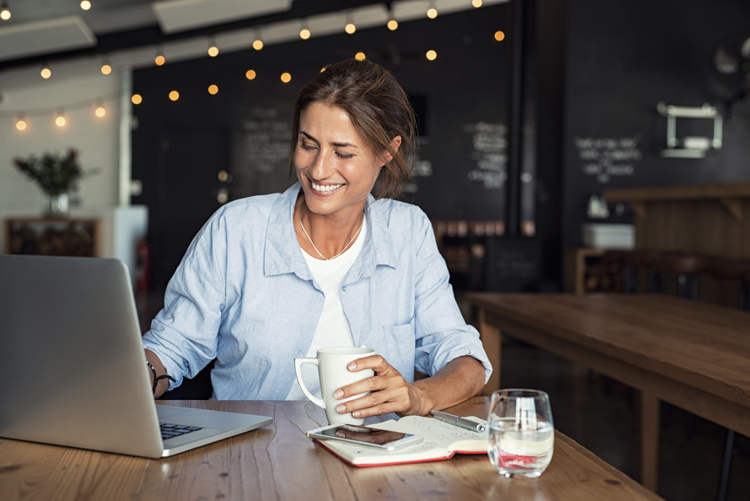 Mature woman working on laptop Smiling woman sitting in cafeteria holding coffee mug and working on laptop. Businesswoman checking email on laptop. Beautiful middle aged woman smiling and using laptop at cafe while drinking a cup of tea.
